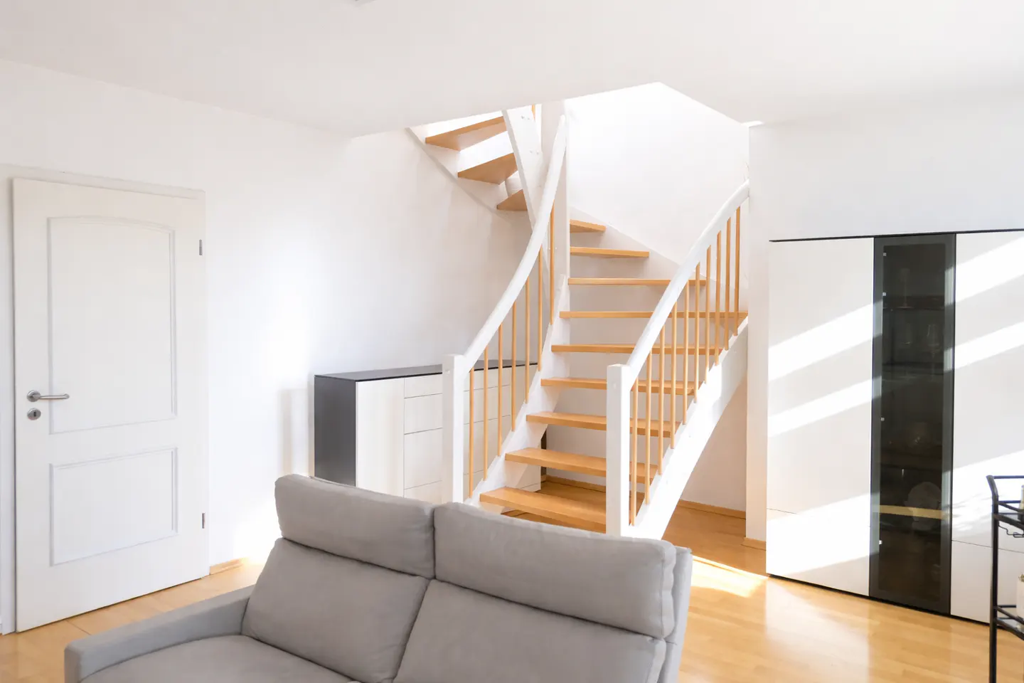 Bright living room with a gray sofa, white door, and a wooden staircase with white railings. A modern cabinet stands against the white wall.