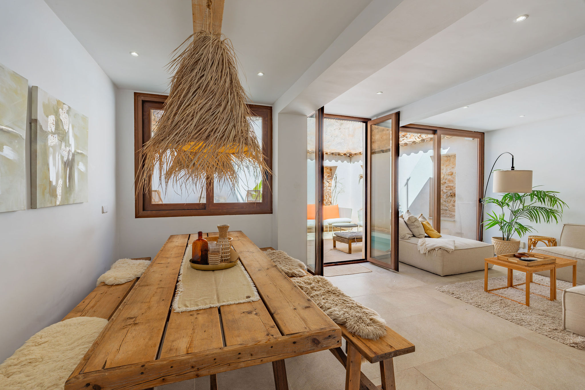 Bright, modern interior with a rustic wood table, benches, and straw chandelier. Glass doors open to an outdoor lounge area.