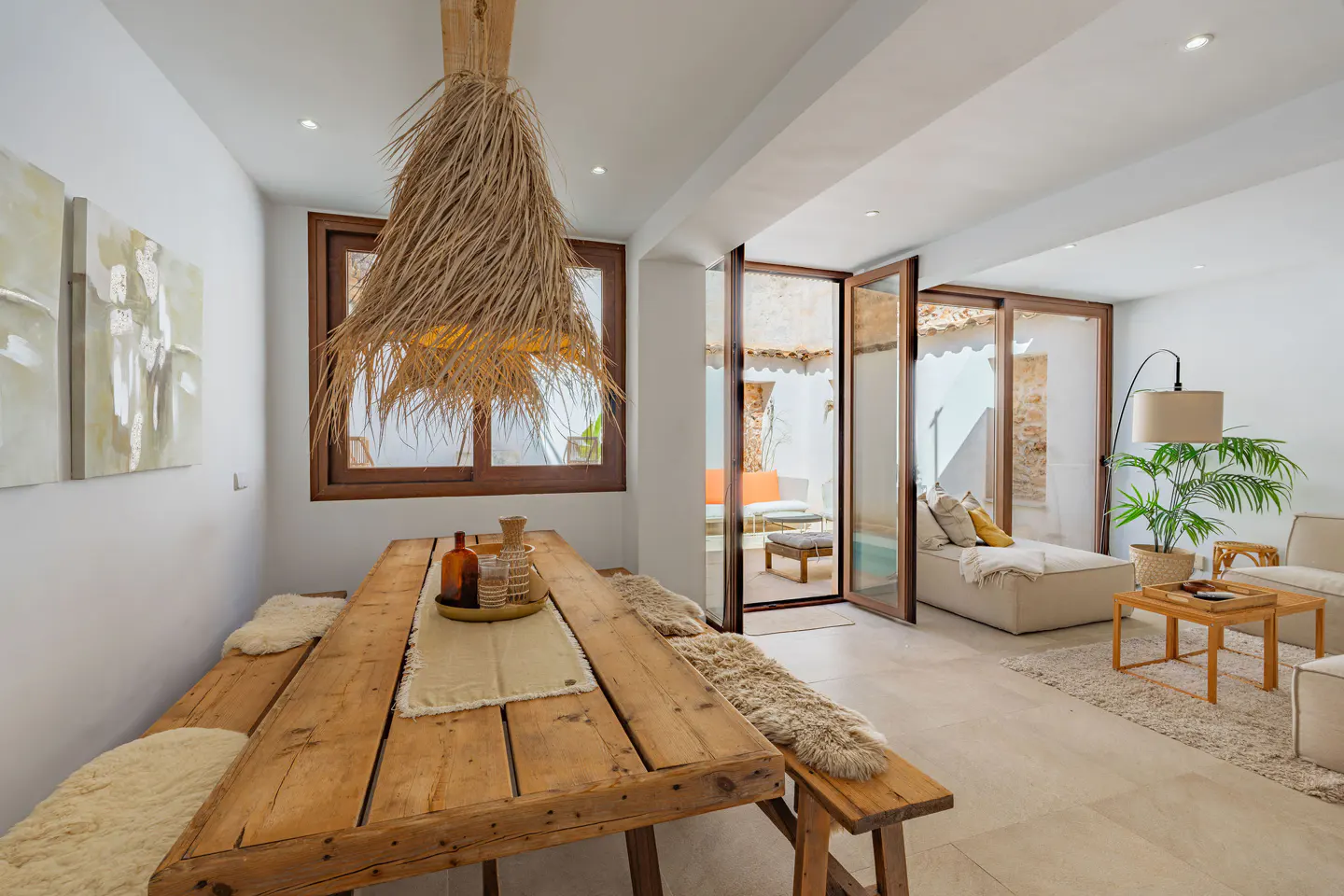 Bright, modern interior with a rustic wood table, benches, and straw chandelier. Glass doors open to an outdoor lounge area.