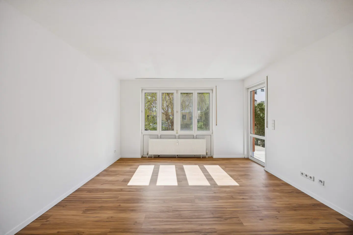 Bright, empty room with wood floors, white walls, and a large window. Sunlight streams in, creating stripes on the floor. A radiator sits under the window.
