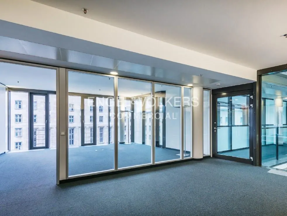 Empty office space with blue carpet, glass walls, and a view of buildings outside. "ENGEL & VOLKERS COMMERCIAL" is printed on the glass.