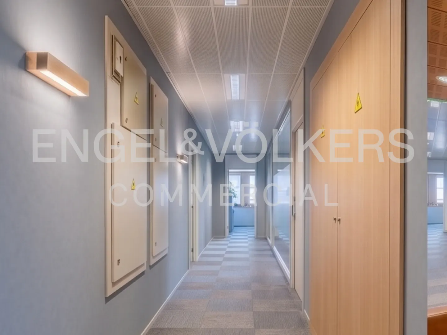 Modern office hallway with grey walls, light wood doors, checkered carpet, and recessed ceiling lighting.