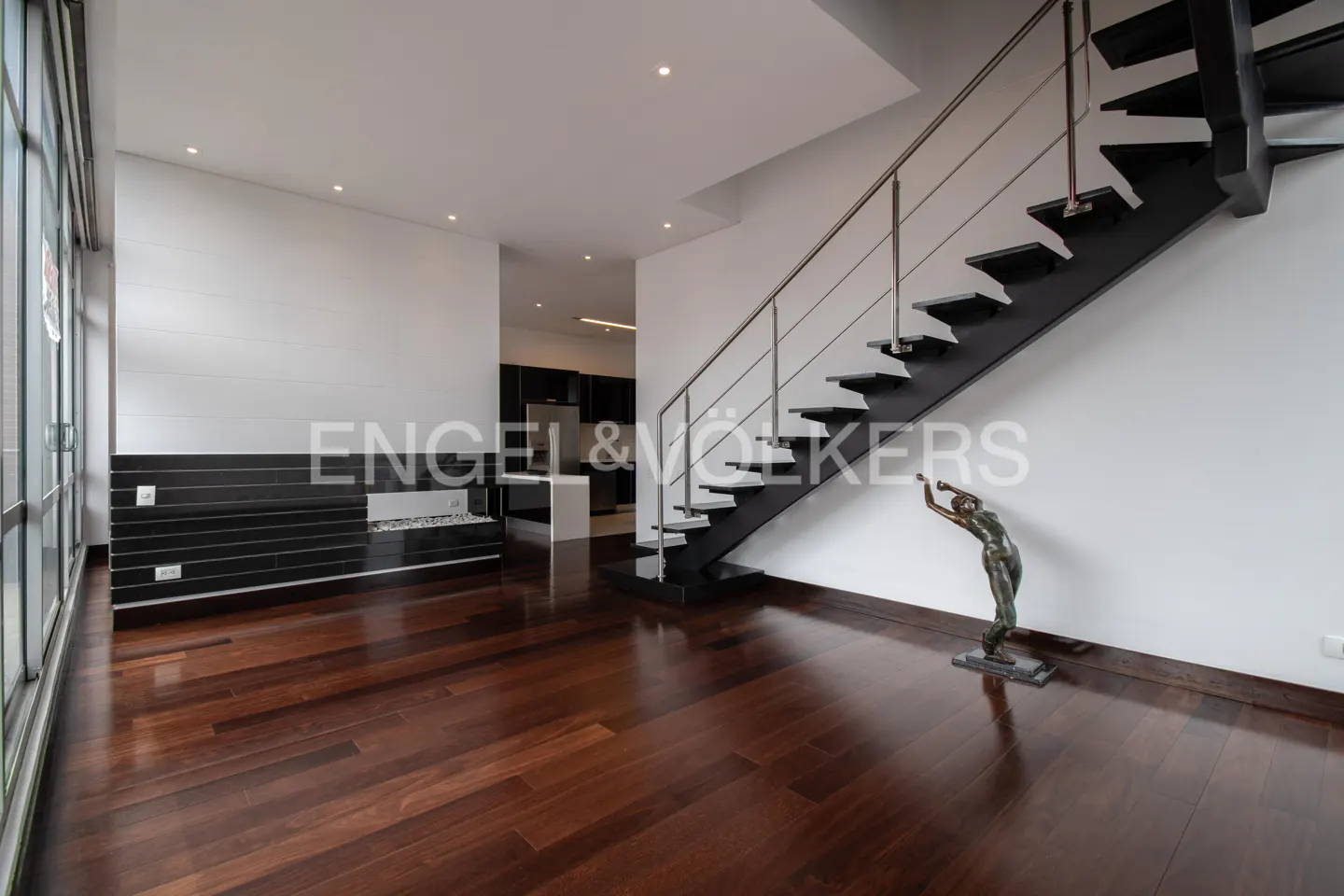Modern living room with dark wood floors, black stairs with steel railing, and a black fireplace. A bronze statue stands near the stairs.