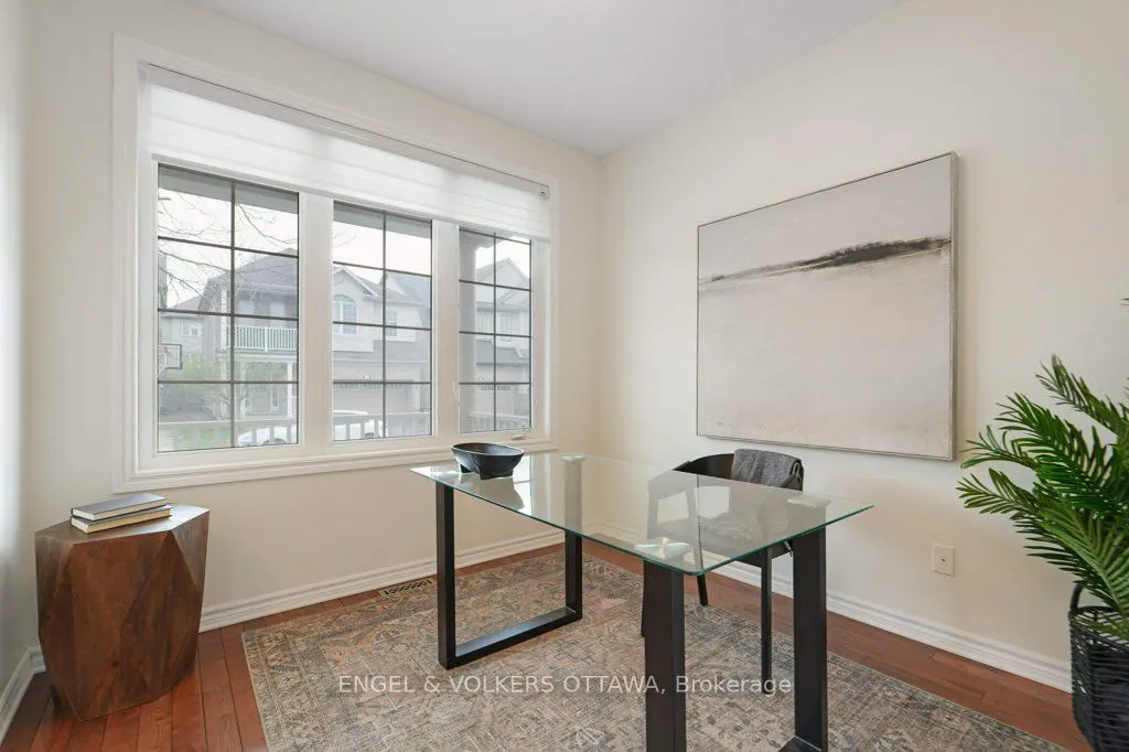 Bright home office with a glass desk, black frame, and a black chair. A large window overlooks a suburban street. Neutral tones and wood floors.