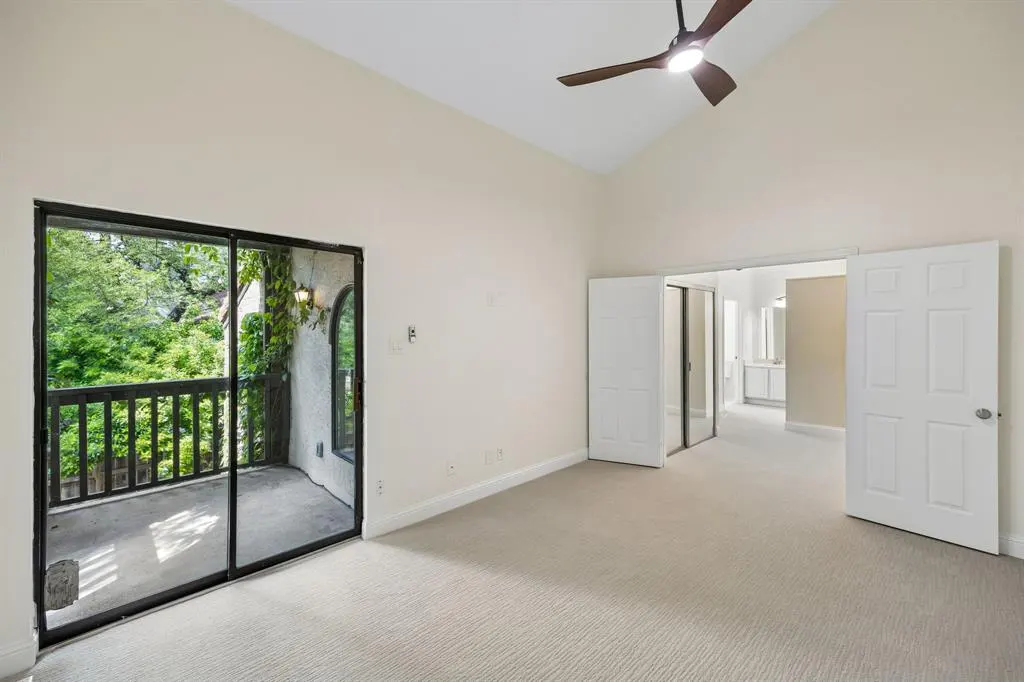 A bright, empty bedroom with beige carpet, white walls, and a ceiling fan. Sliding glass doors lead to a balcony with green trees.