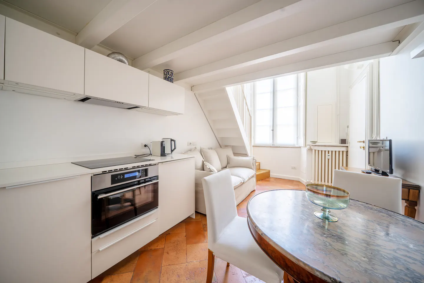 Modern white kitchen with stainless steel oven, dining area with marble table, and a cozy sofa under a staircase.