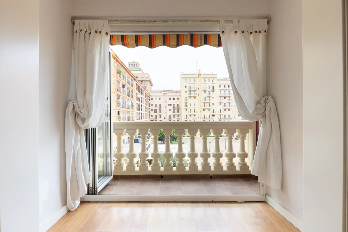 Open balcony door with white curtains, showing a city view with beige buildings and a striped awning.