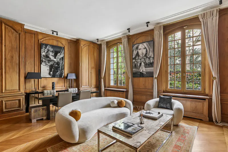 Living room with wood paneling, parquet floors, and large windows. A curved white sofa faces a marble coffee table. Artwork adorns the walls.