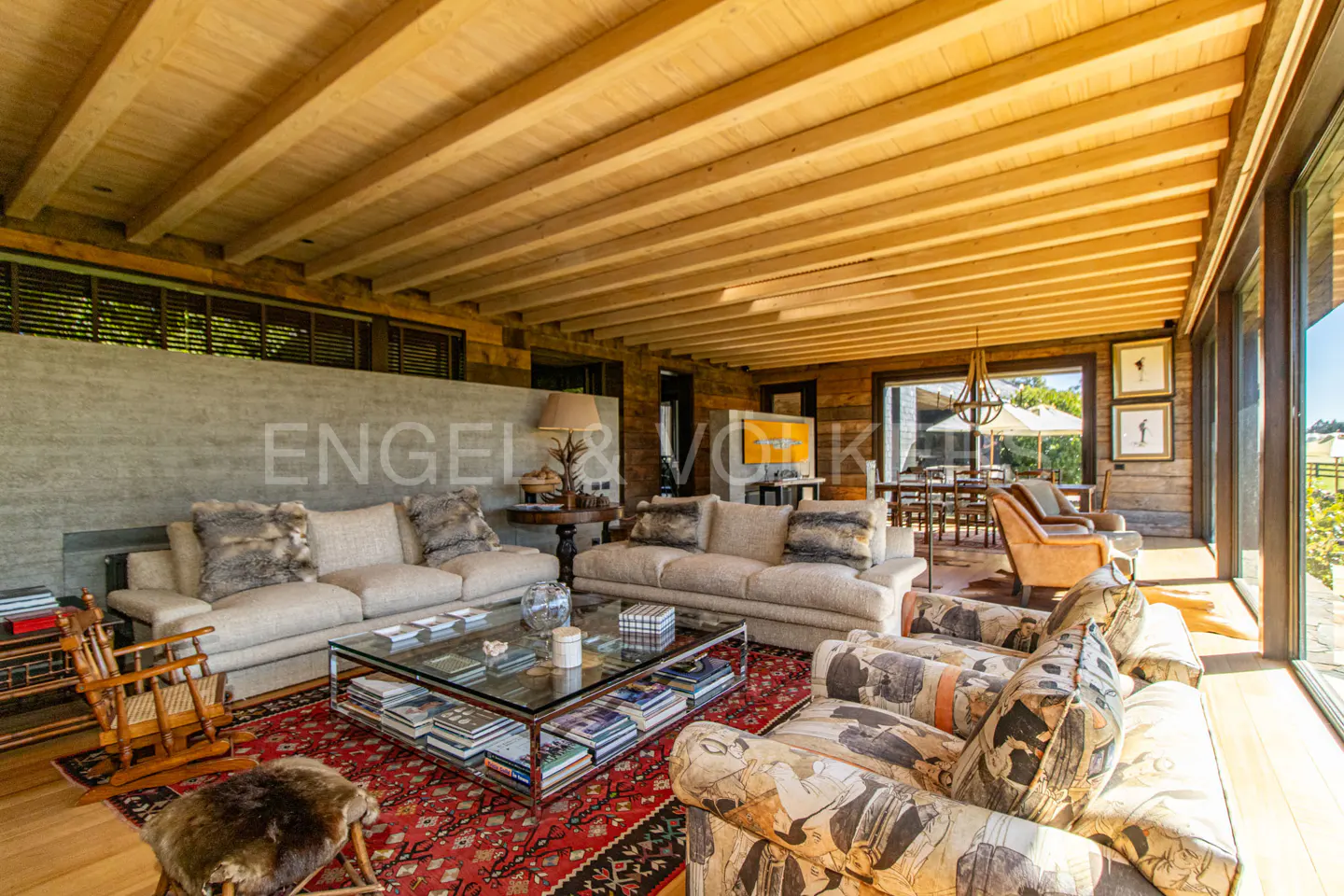A spacious living room with wood ceilings, sofas, a glass coffee table, and a red patterned rug. Large windows offer natural light.