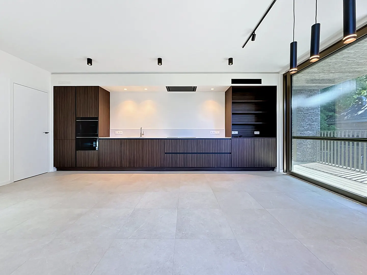 A modern kitchen with dark wood cabinets, stainless steel appliances, and light gray tile flooring. Large windows offer natural light.
