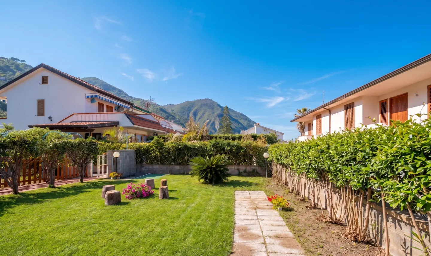 A sunny backyard with green grass, a stone path, and trimmed hedges between two white houses. Mountains are visible in the background.