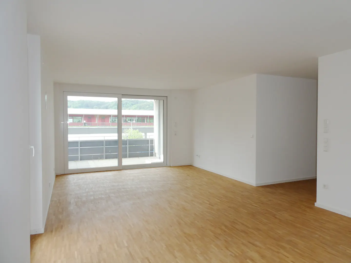 Bright, empty room with light wood floors and white walls. Sliding glass doors lead to a balcony with a view of a building and trees.
