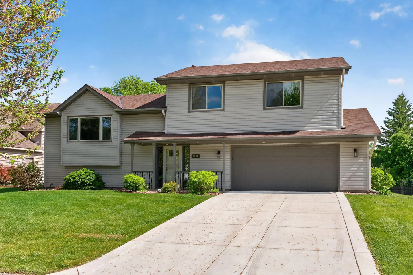 Two-story house with gray siding, brown roof, and a gray garage door on a sunny day. A concrete driveway leads to the garage.