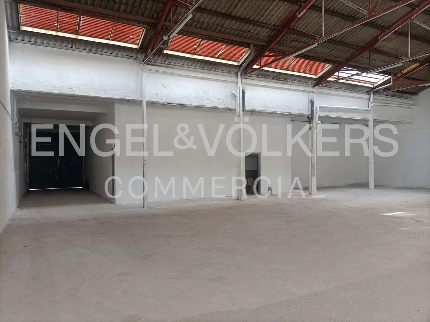 Empty warehouse interior with concrete floor, white walls, and skylights in the red metal roof. "Engel & Volkers Commercial" is overlaid.