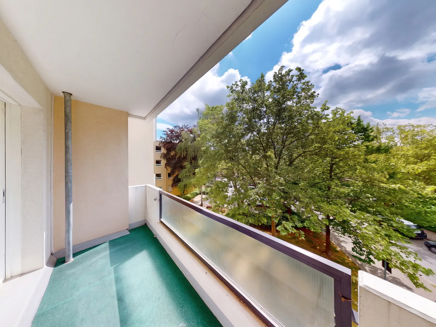 A balcony with a green floor and frosted glass railing overlooking lush green trees under a blue sky with white clouds.