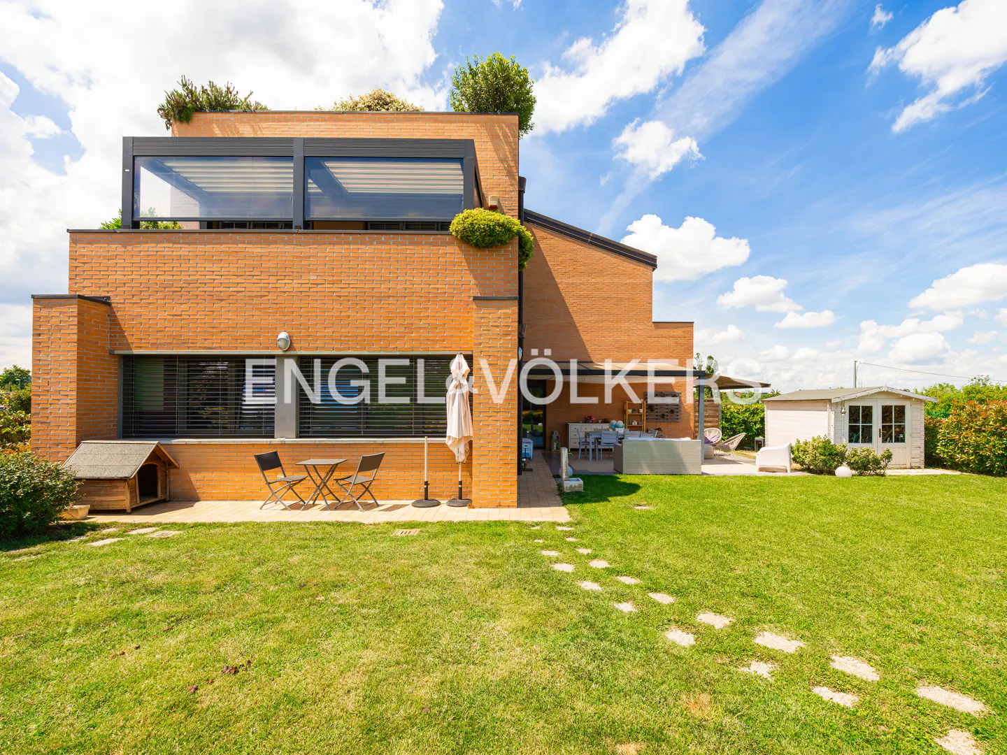 Backyard view of a modern brick house with a green lawn, patio furniture, and a small shed under a blue sky with clouds.
