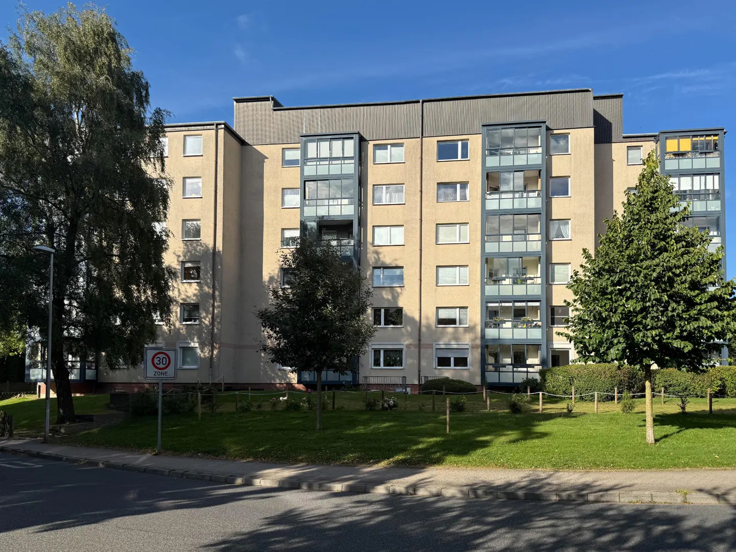 Exterior view of a multi-story apartment building with beige walls and gray balconies, surrounded by green lawns and trees.