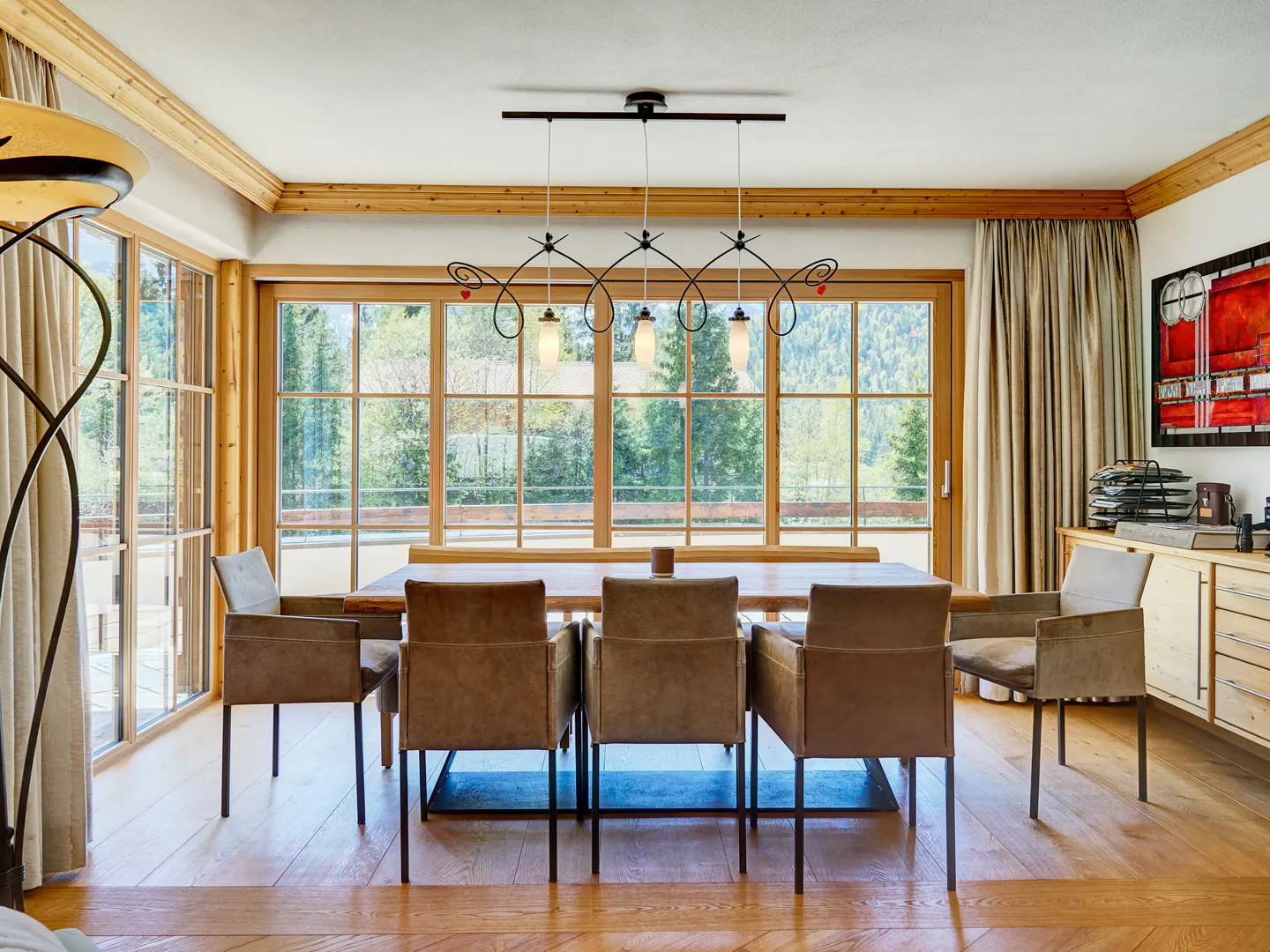 Dining room with wood floors, table, and chairs. Large windows overlook a forest. A modern chandelier hangs above the table.