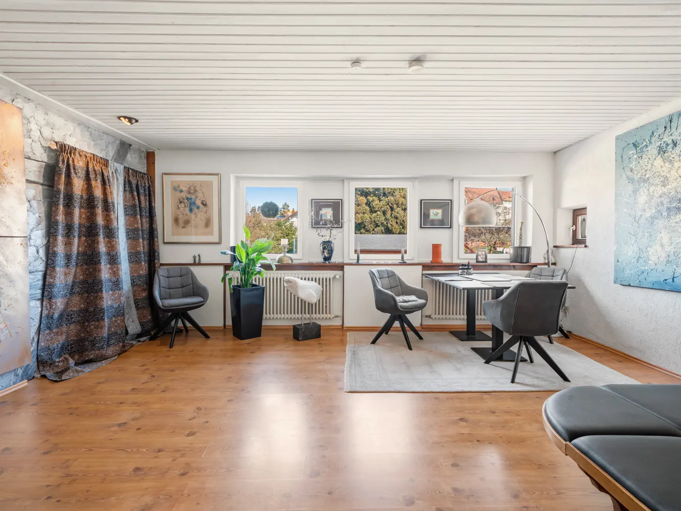 Bright room with wood floors, white walls, and multiple windows. Gray chairs surround a black table on a white rug. Art and plants decorate the space.
