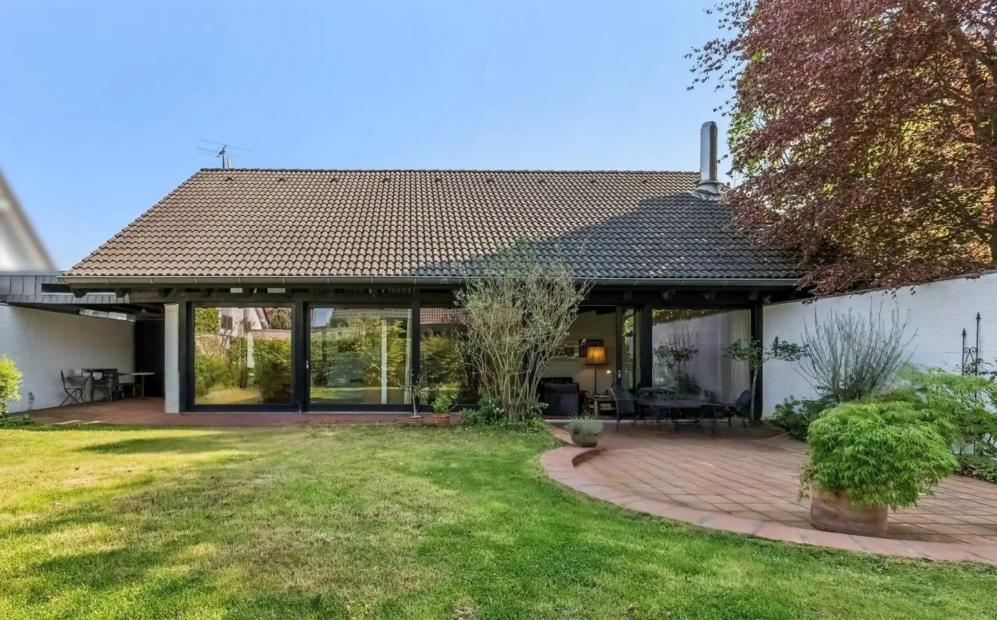 Backyard view of a single-story house with a brown tiled roof, black framed windows, and a brick patio with outdoor furniture.