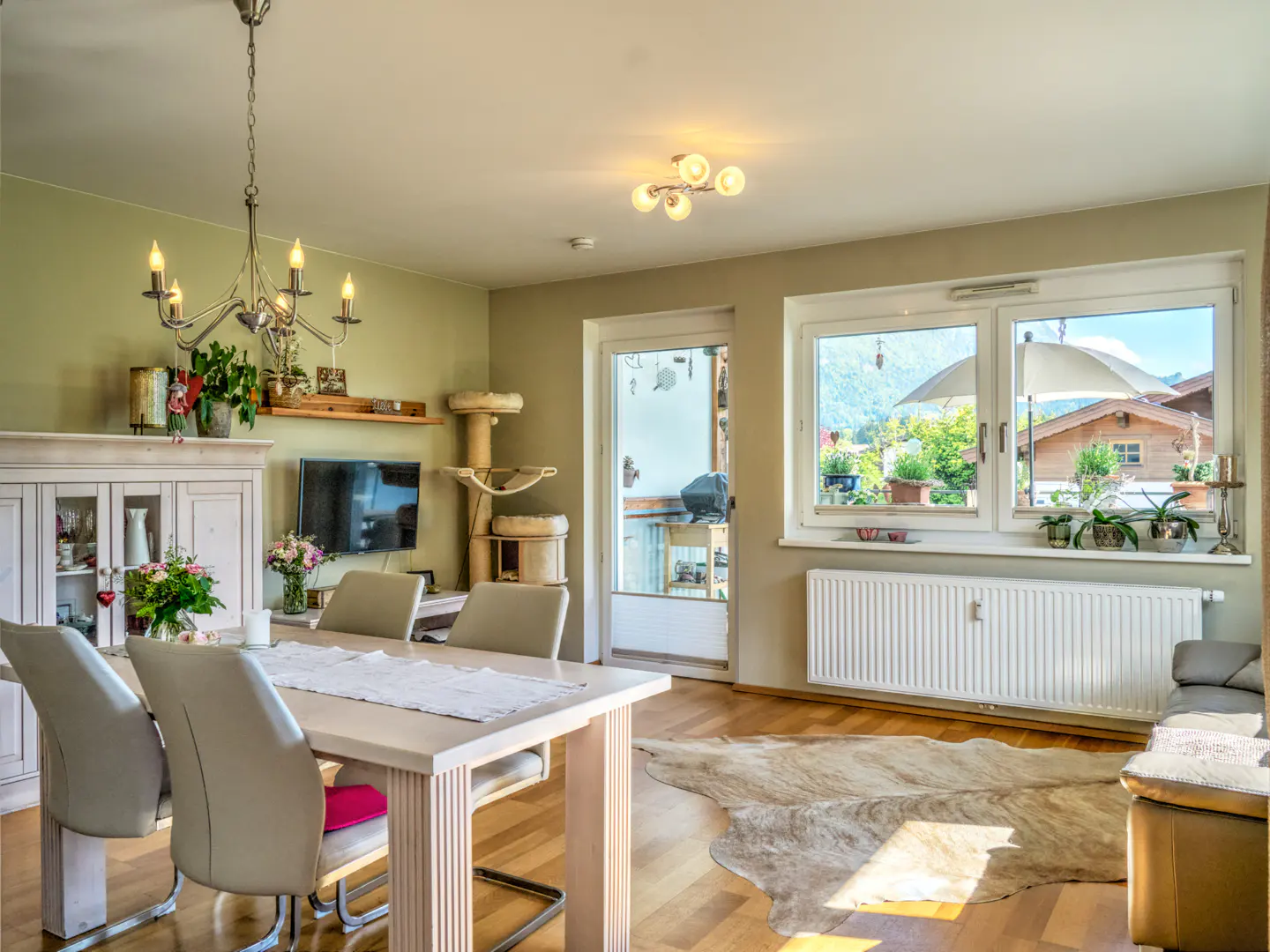A bright living room with a dining table, chairs, a TV, and a cat tree. Outside the window, there are houses and trees.