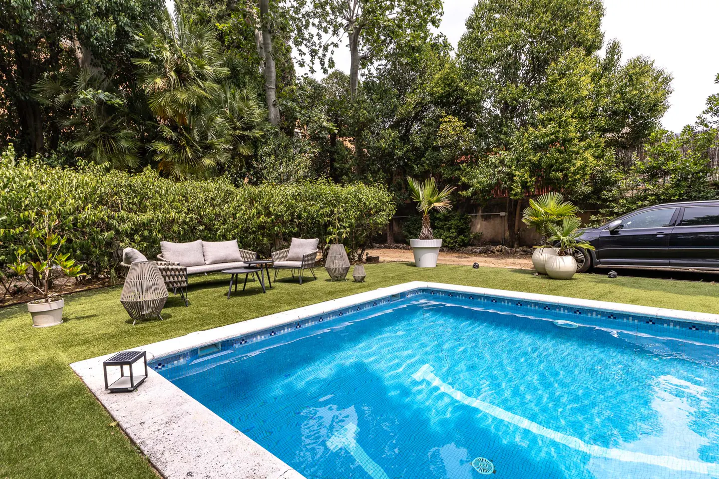 A backyard swimming pool with blue water, patio furniture on artificial grass, and lush green trees in the background.