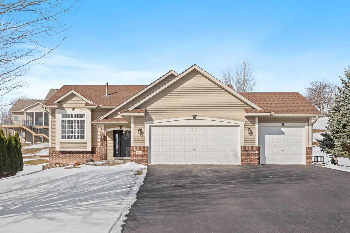 Beige siding house with a brown roof, white garage doors, and a black front door on a sunny day.