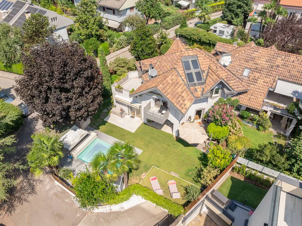 Aerial view of a white house with a brown tile roof, a pool, and a green lawn surrounded by trees.