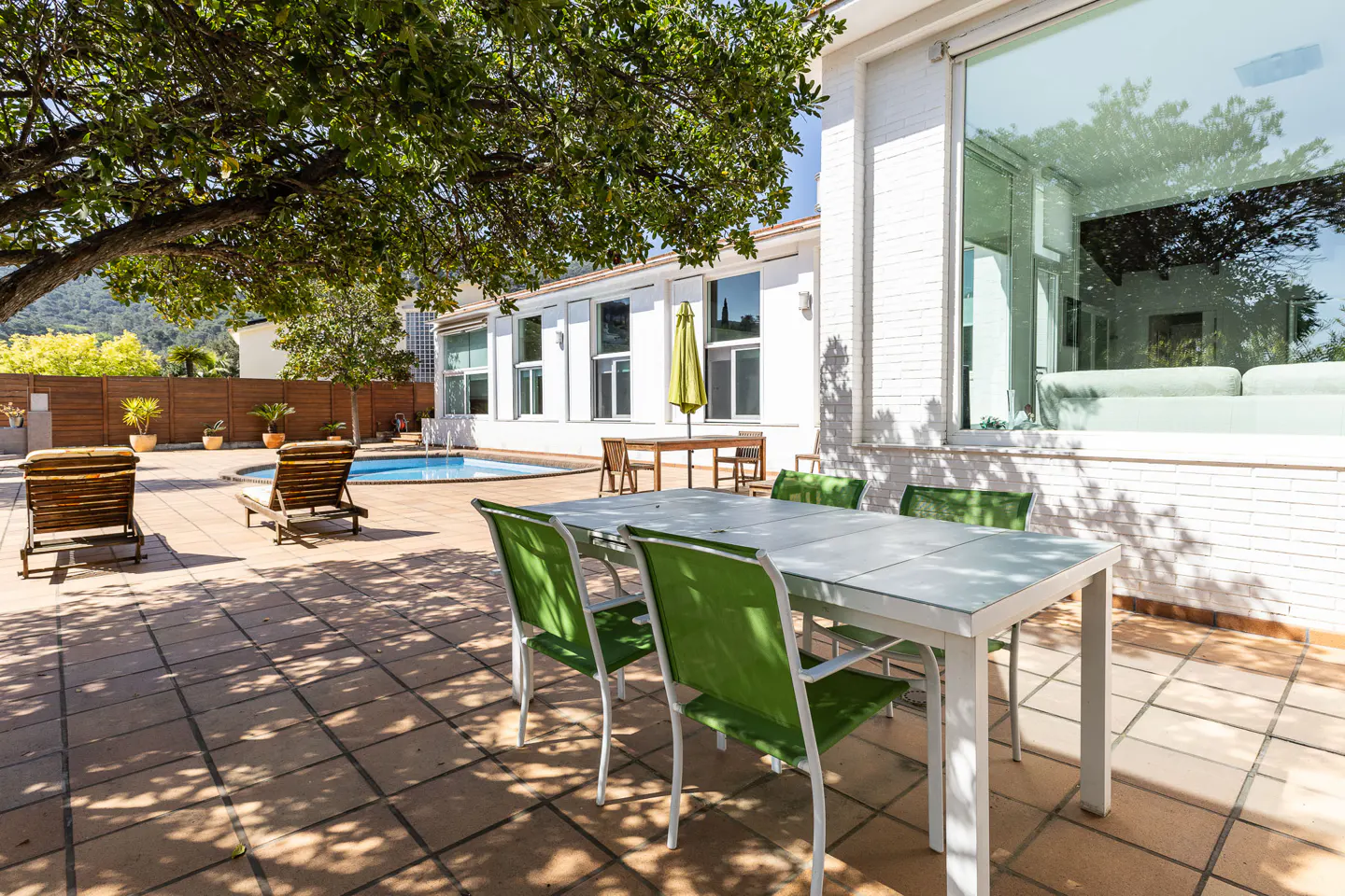 Outdoor patio with a white table, green chairs, lounge chairs, and a pool. A large tree provides shade.