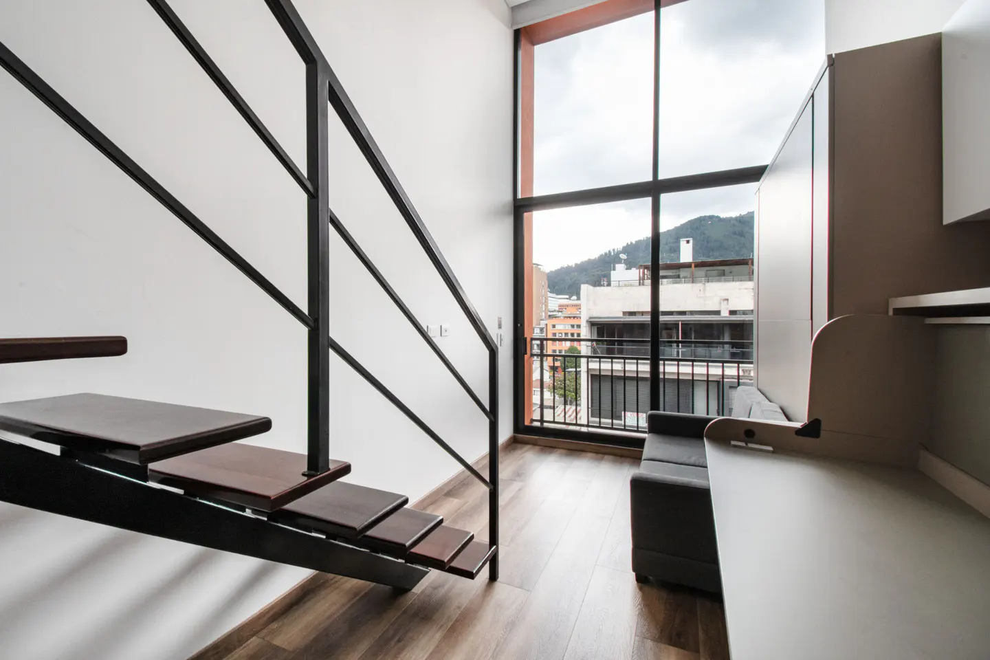 Interior view of a modern apartment with a black metal staircase, large window with city view, and a gray sofa.