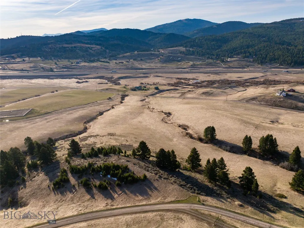 Aerial view of a Montana landscape with brown fields, green trees, and mountains in the background under a blue sky.