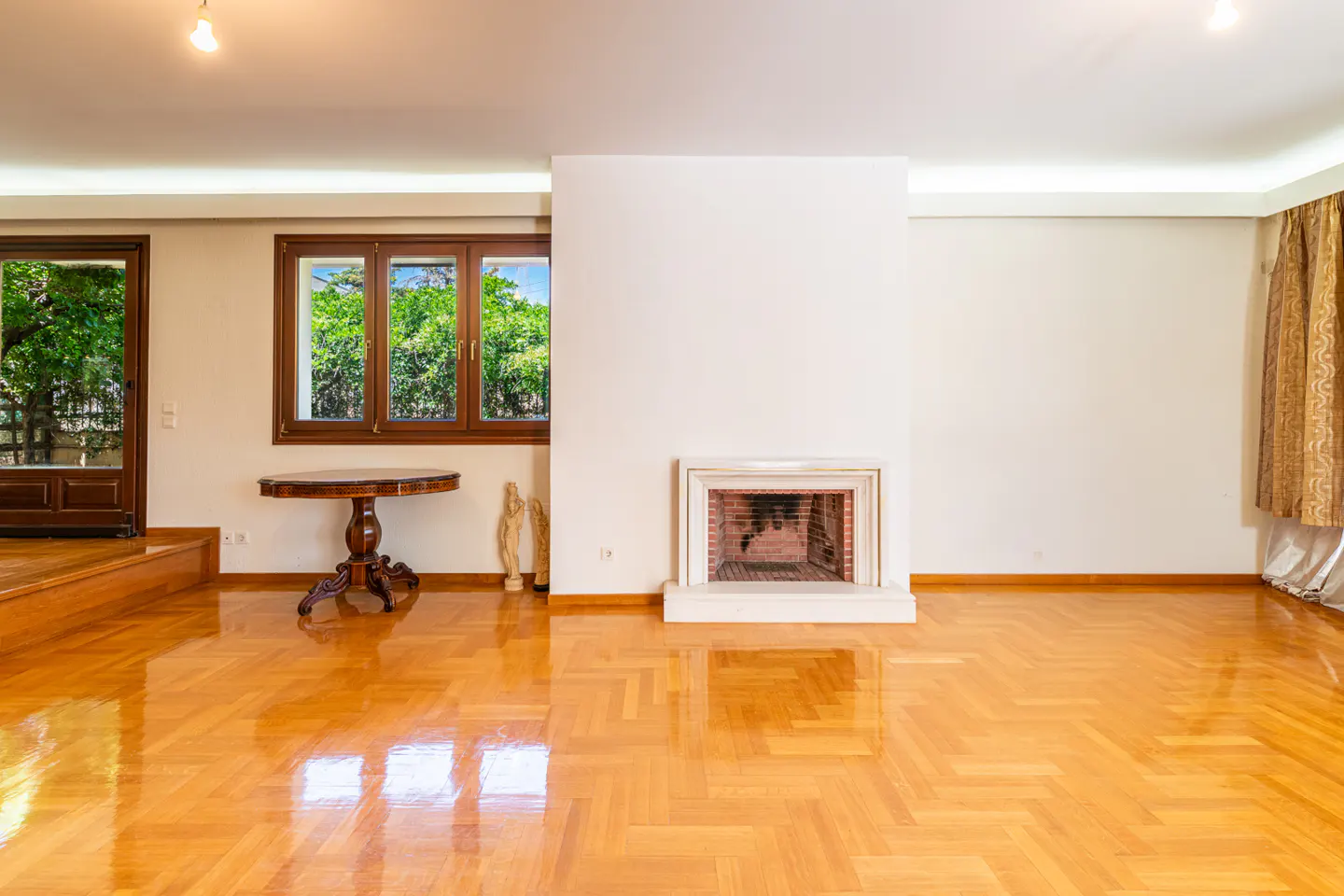 Spacious living room with polished herringbone wood floors, a central fireplace, a wooden table, and garden views.