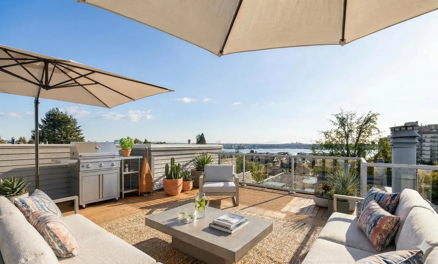 Rooftop patio with outdoor furniture, grill, and city view. Two large umbrellas provide shade. The patio has a wooden deck and a concrete coffee table.