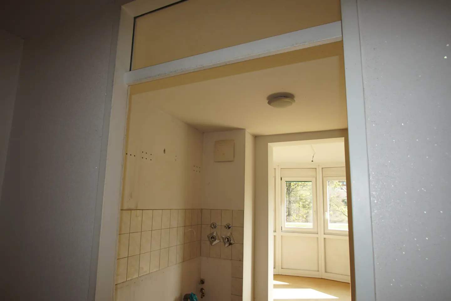 A view into a small, unfinished kitchen with white walls and tile, and a bright sunroom beyond.