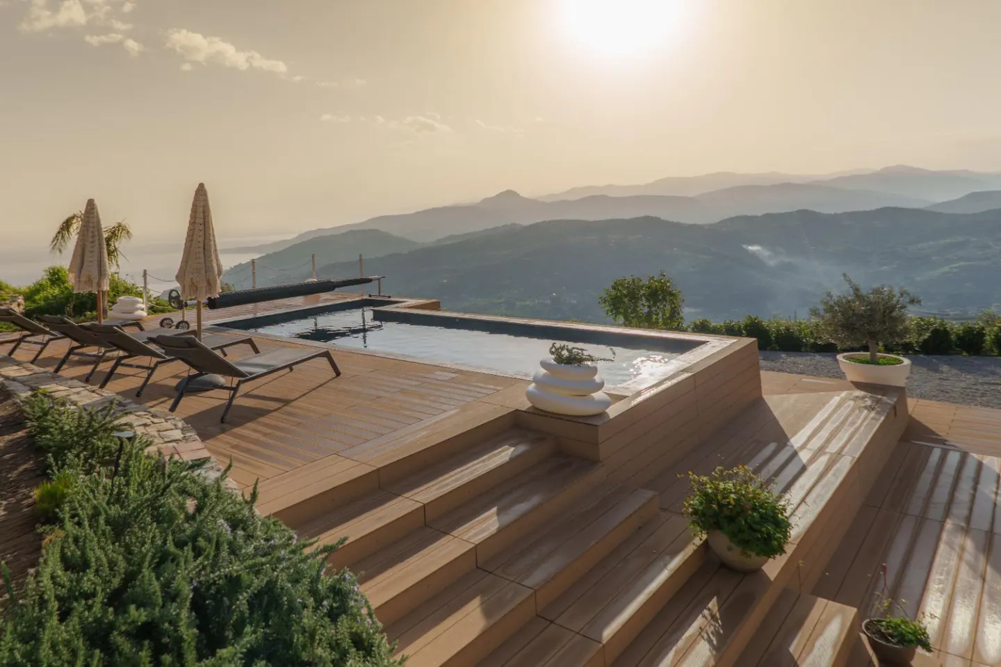 Luxury pool deck with lounge chairs, umbrellas, and mountain views. The pool is surrounded by wooden decking and steps.