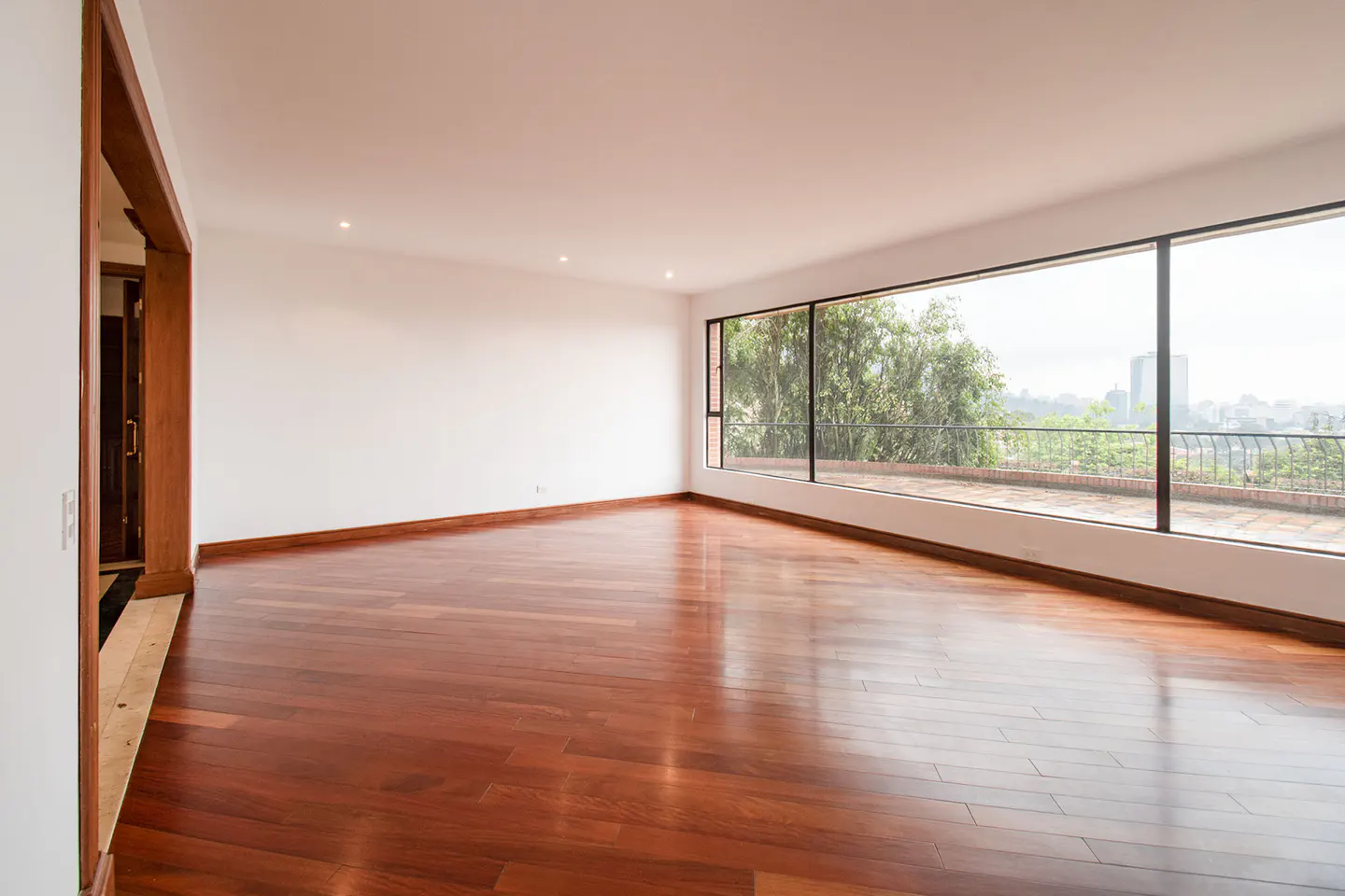 Bright, empty room with hardwood floors and white walls. Large window overlooks a balcony, trees, and city skyline.