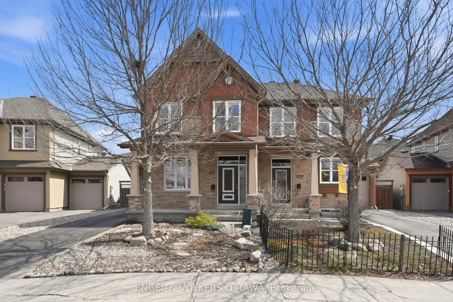 Two-story brick townhouse with a brown roof and black doors, framed by bare trees and a small black fence.