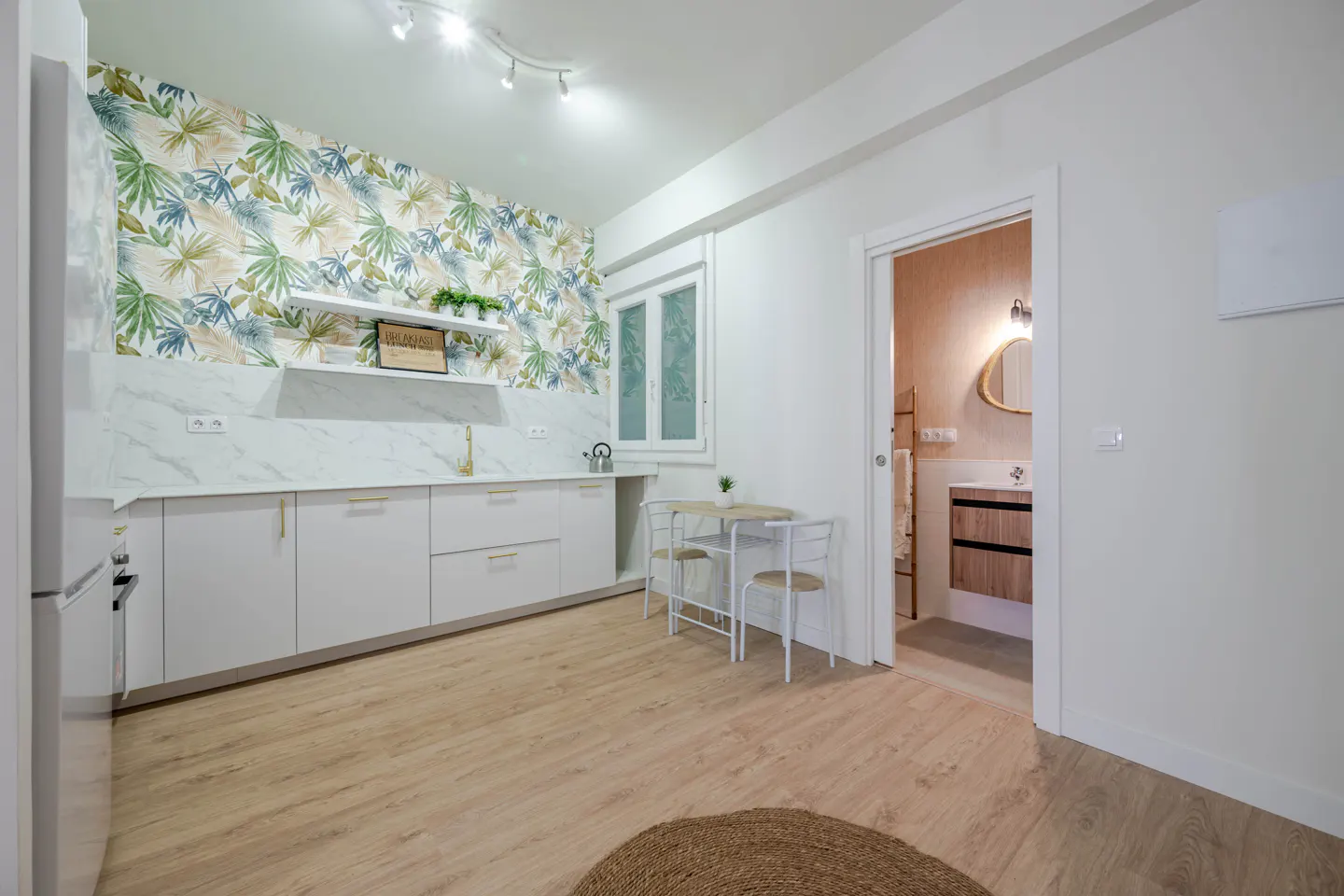 Bright kitchen with white cabinets, marble counters, and tropical wallpaper. A small table and chairs sit near an open bathroom door.