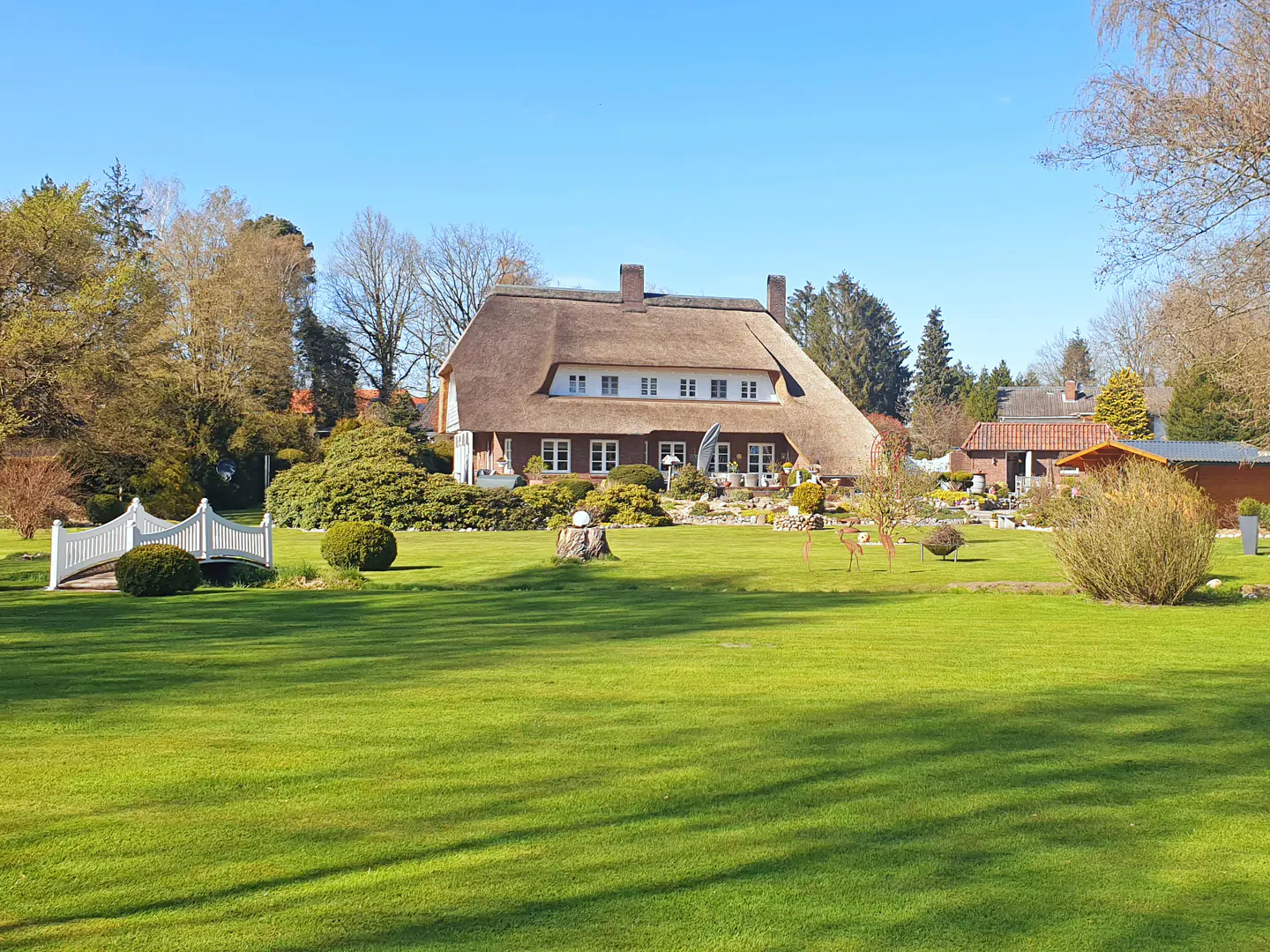 A large house with a thatched roof sits on a green lawn under a blue sky. A white bridge is on the left.