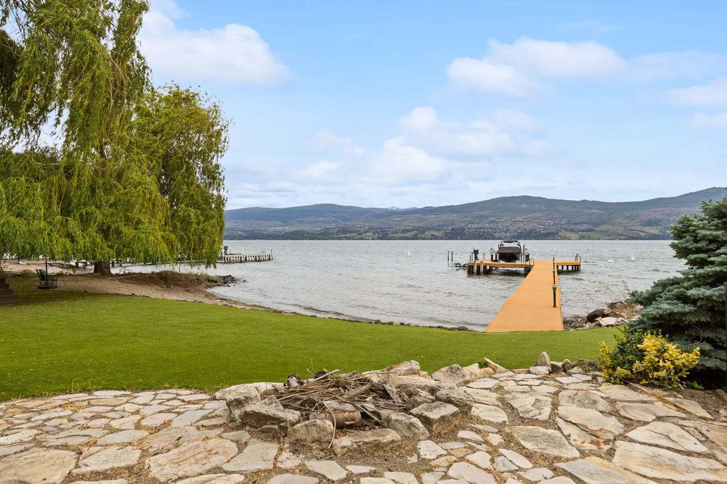 Waterfront property with a stone patio, green lawn, and a wooden dock extending into a lake with mountains in the background.