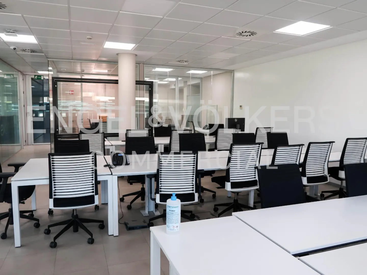 Bright office space with white desks and black chairs. A bottle of hand sanitizer sits on a desk. Glass walls and white ceiling tiles.