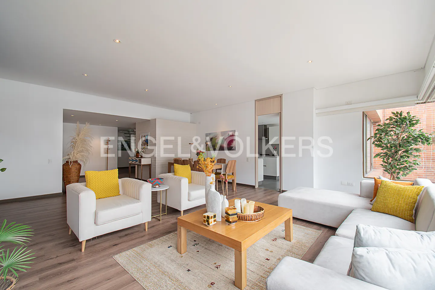 Bright, open living room with white sofas, yellow pillows, and a wooden coffee table on a patterned rug. Dining area and kitchen visible in the background.