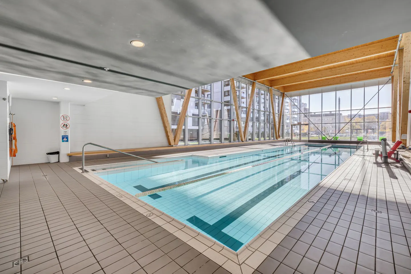 Indoor pool with blue water and lane markers, surrounded by gray tile and a wall of windows with wooden beams.