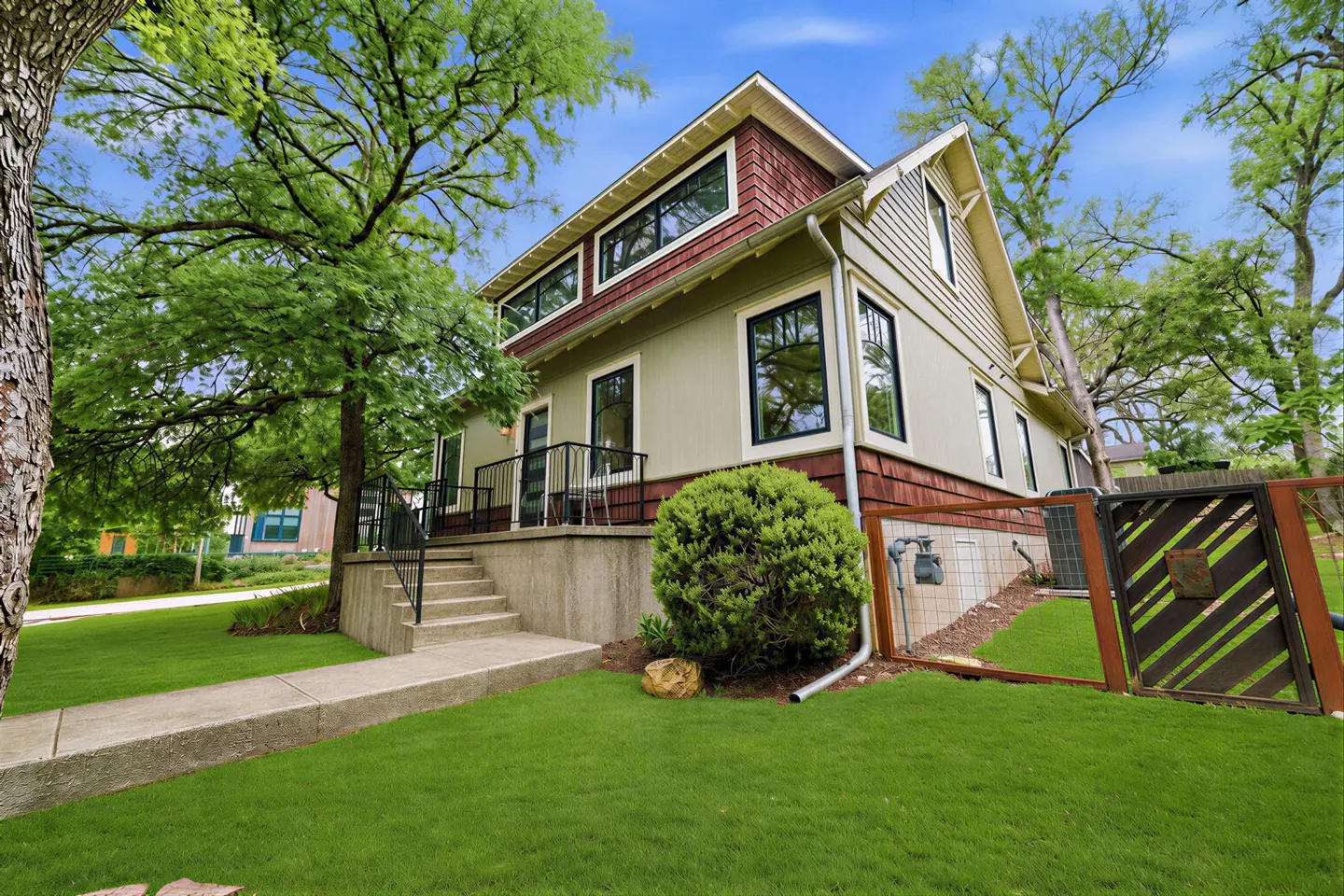 Two-story house with green siding, red shingle accents, black-framed windows, and a concrete porch with black railings. Green lawn and trees surround the house.
