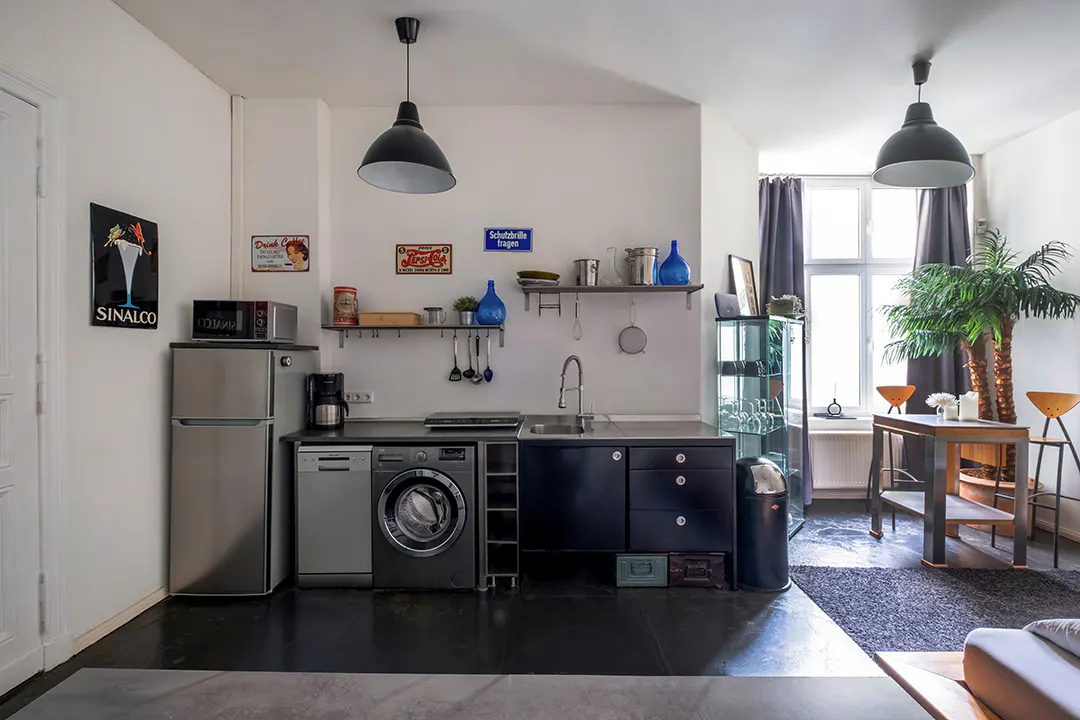 A modern kitchen with stainless steel appliances, dark cabinets, and open shelving with blue accents. A dining area with a palm tree is visible.