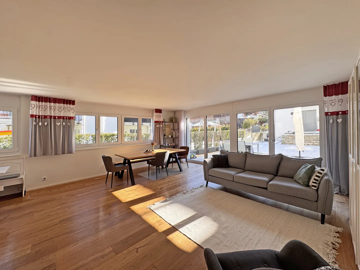 Bright living room with wood floors, a gray sofa, dining table, and large windows with heart-patterned curtains.
