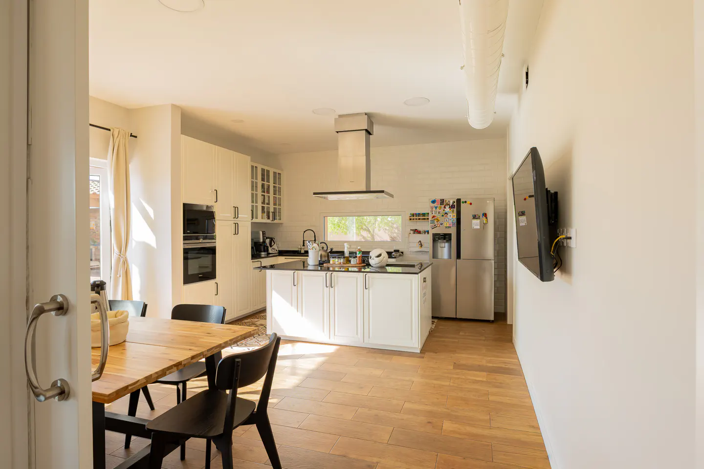 Bright, modern kitchen with white cabinets, stainless steel appliances, and a wood dining table with black chairs. A TV is mounted on the wall.