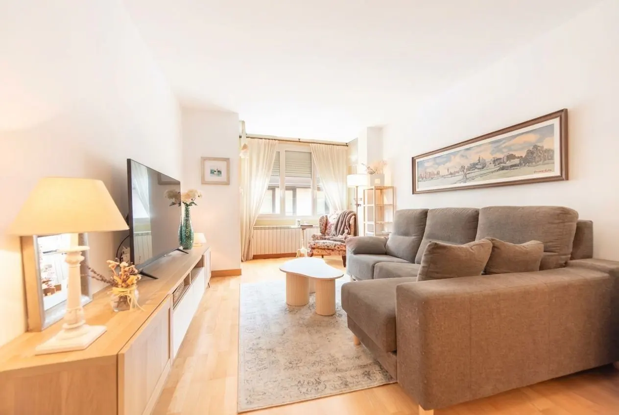 Bright living room with a brown sofa, light wood floors, and a TV on a light wood console. A window with white curtains is in the background.
