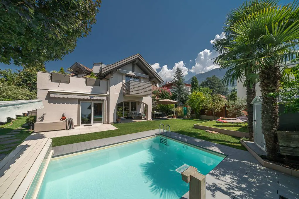 A backyard with a pool, a white house, green grass, and palm trees under a blue sky.