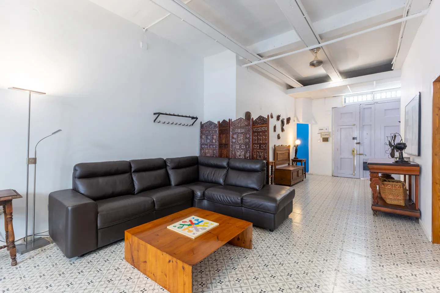 Living room with a brown leather sectional sofa, wooden coffee table, and patterned tile floor. A carved screen and vintage fan add character.